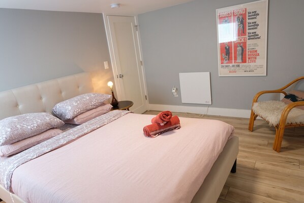 Un lit bien fait, avec une literie rose et une serviette enroulée, une chaise en bois dotée d’un coussin moelleux et une affiche de film accrochée au mur.