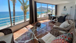 A spacious balcony with a view of the sea and a mountain, furnished with wicker furniture and a glass coffee table.