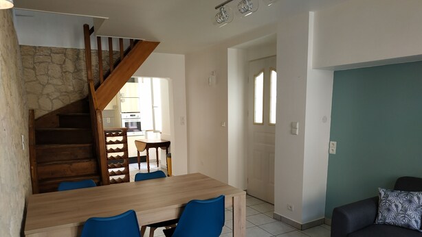Zona pranzo con tavolo in legno e sedie blu, una scala con ringhiere in legno, una cucina sullo sfondo e un divano scuro con un cuscino decorato.