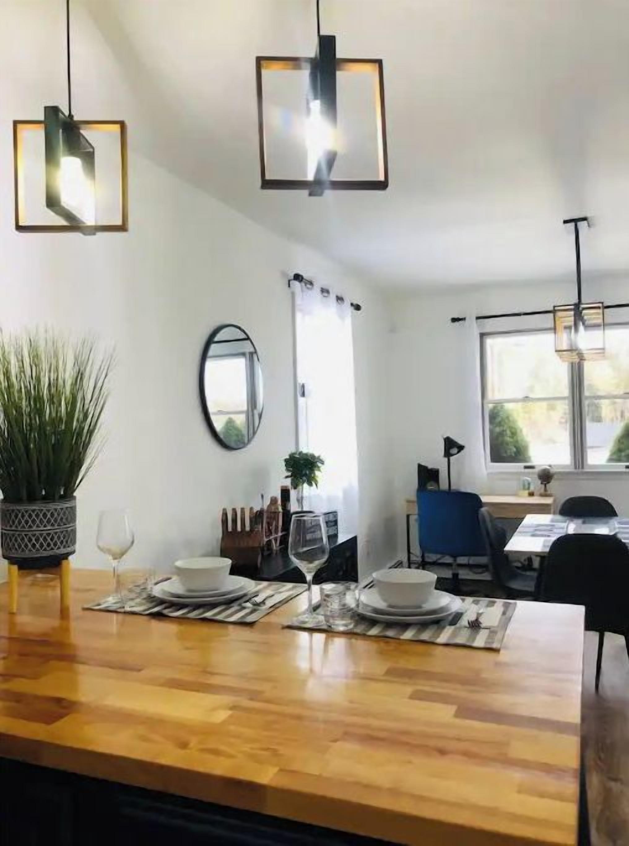 A modern dining area with a wooden table set for two, two pendant lights, a potted plant, and a mirror on the wall.
