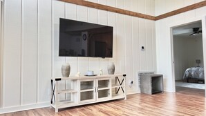 Quarto com piso de madeira, paredes brancas com painéis e uma televisão de tela plana fixada acima de um console de madeira com portas de vidro. Uma cama está no quarto vizinho.