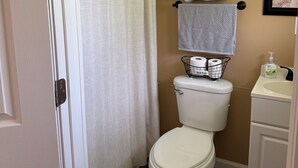 A bathroom with a toilet, a shower curtain, a towel rack with towels, a toilet paper holder, and a sink with a soap dispenser.