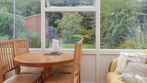 Une pièce avec une table et des chaises en bois, une fenêtre donnant sur un paysage verdoyant et un canapé jaune agrémenté de coussins.