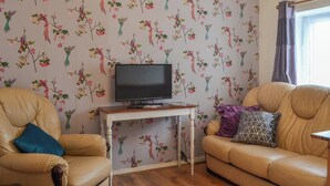 Un salon avec un papier peint à motifs floraux, un téléviseur posé sur une table en bois, un canapé en cuir beige et une fenêtre avec des rideaux.