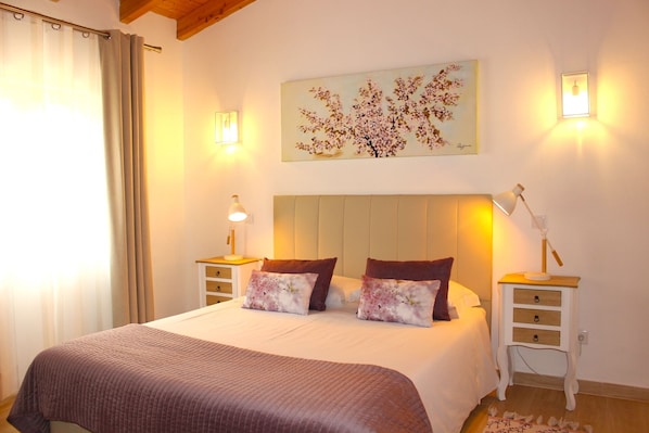 Une chambre à coucher avec un grand lit, deux tables de chevet avec des lampes, un tableau accroché au mur et une fenêtre avec des rideaux.