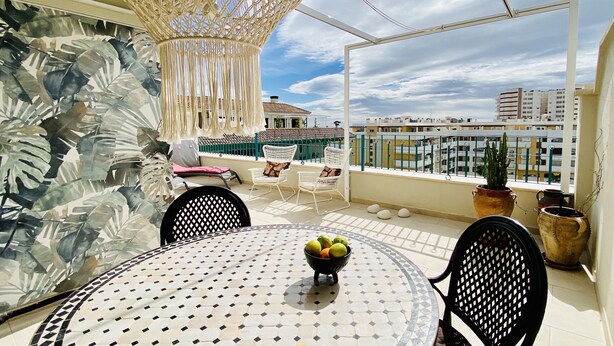 Un balcon avec une table ronde, des chaises, un luminaire suspendu et une vue sur les bâtiments en arrière-plan.
