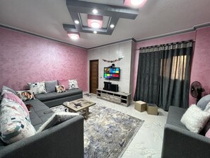 Uma sala de estar com um sofá cinzento, uma mesa de centro, uma TV na parede e um tapete estampado.