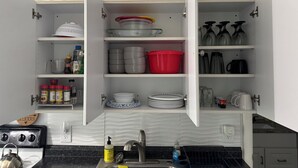 Una cocina con armarios blancos, una estufa y un lavabo. Se observan diversos utensilios de cocina como tazas, platos y una olla roja.