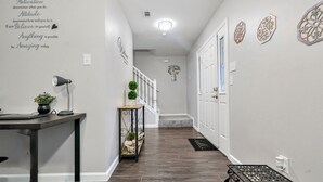 Un couloir avec un plancher en bois foncé, des murs blancs et un luminaire au plafond. On y trouve une petite table sur laquelle sont posés une lampe et une plante, ainsi qu’un mur décoratif orné de motifs et de texte.