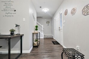 Un couloir avec un plancher en bois foncé, des murs blancs et un luminaire au plafond. On y trouve une petite table sur laquelle sont posés une lampe et une plante, ainsi qu’un mur décoratif orné de motifs et de texte.