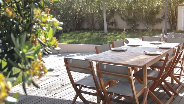 Een terras buiten met een houten tafel en stoelen, omringd door groen en een beschaduwd gedeelte.
