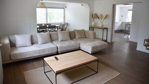 Een moderne woonkamer met een hoekbank, een salontafel en een eethoek, zichtbaar door een boog.