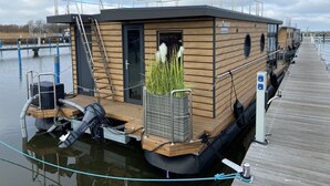 Ein Hausboot an einem Steg, mit Holzaußenverkleidung, einer Leiter und einem kleinen Gartenbereich mit Pflanzen.