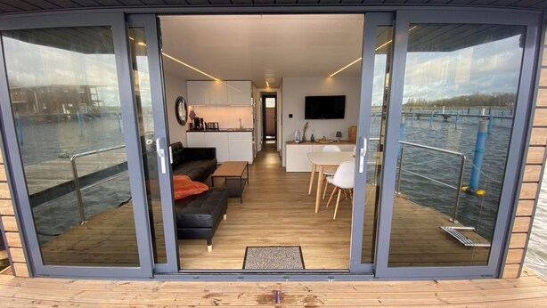 Ein modernes Interieur mit Küche, Essbereich und Sitzgelegenheiten, mit großen Glasüren, die auf eine Terrasse mit Blick auf das Wasser führen.