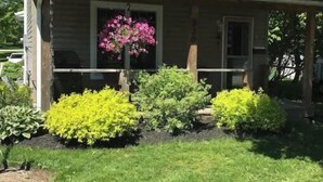 A two-story house with a front porch, a small garden with various shrubs and a flower bed, and a clear blue sky. - Shallow Lake Retreat (Shallow Lake)