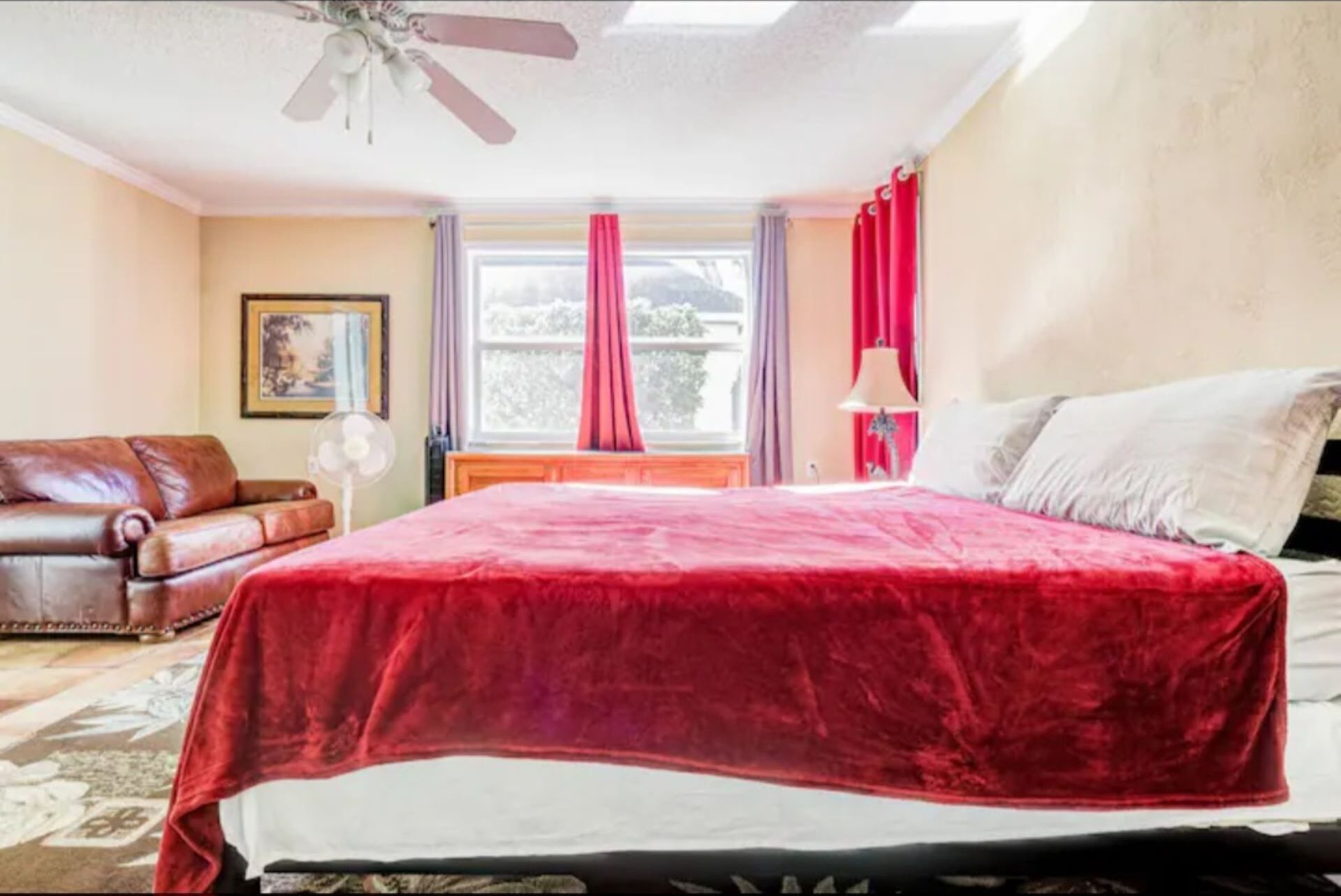 A bedroom with a large bed, a ceiling fan, a window with curtains, a framed picture on the wall, and a brown leather sofa.