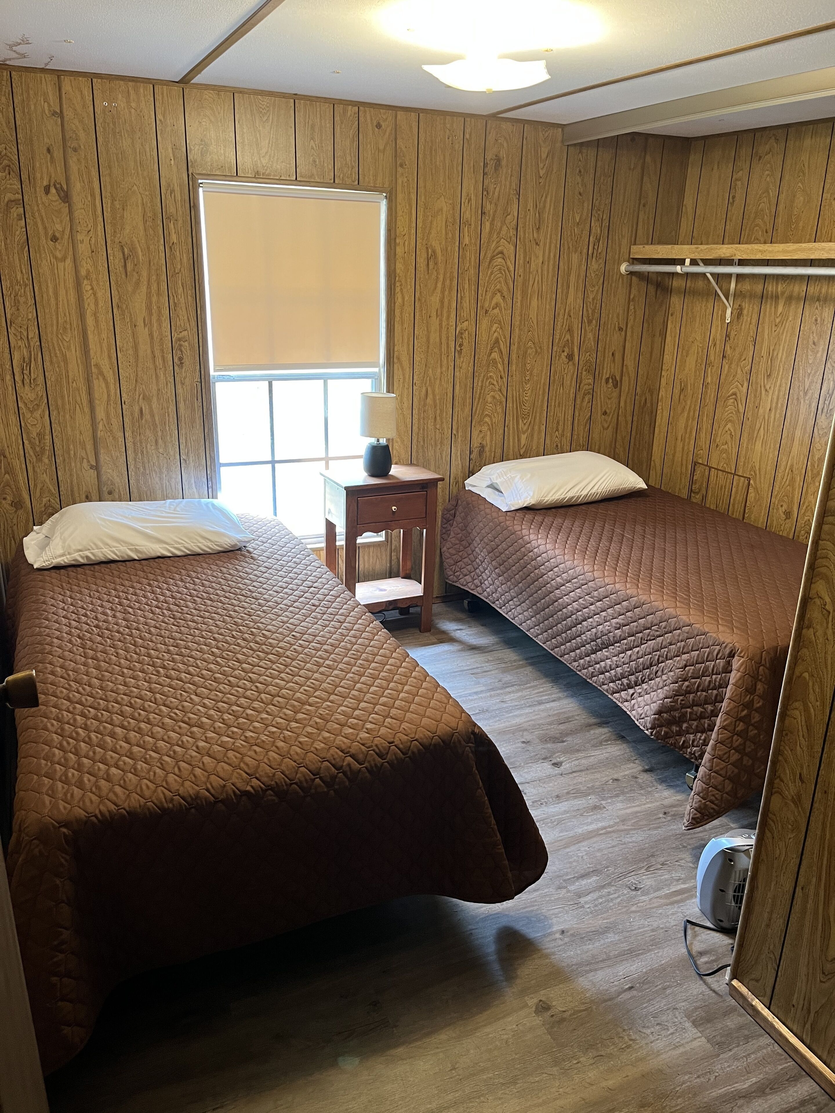 A room with two beds, a small table with a lamp, and a window with blinds.