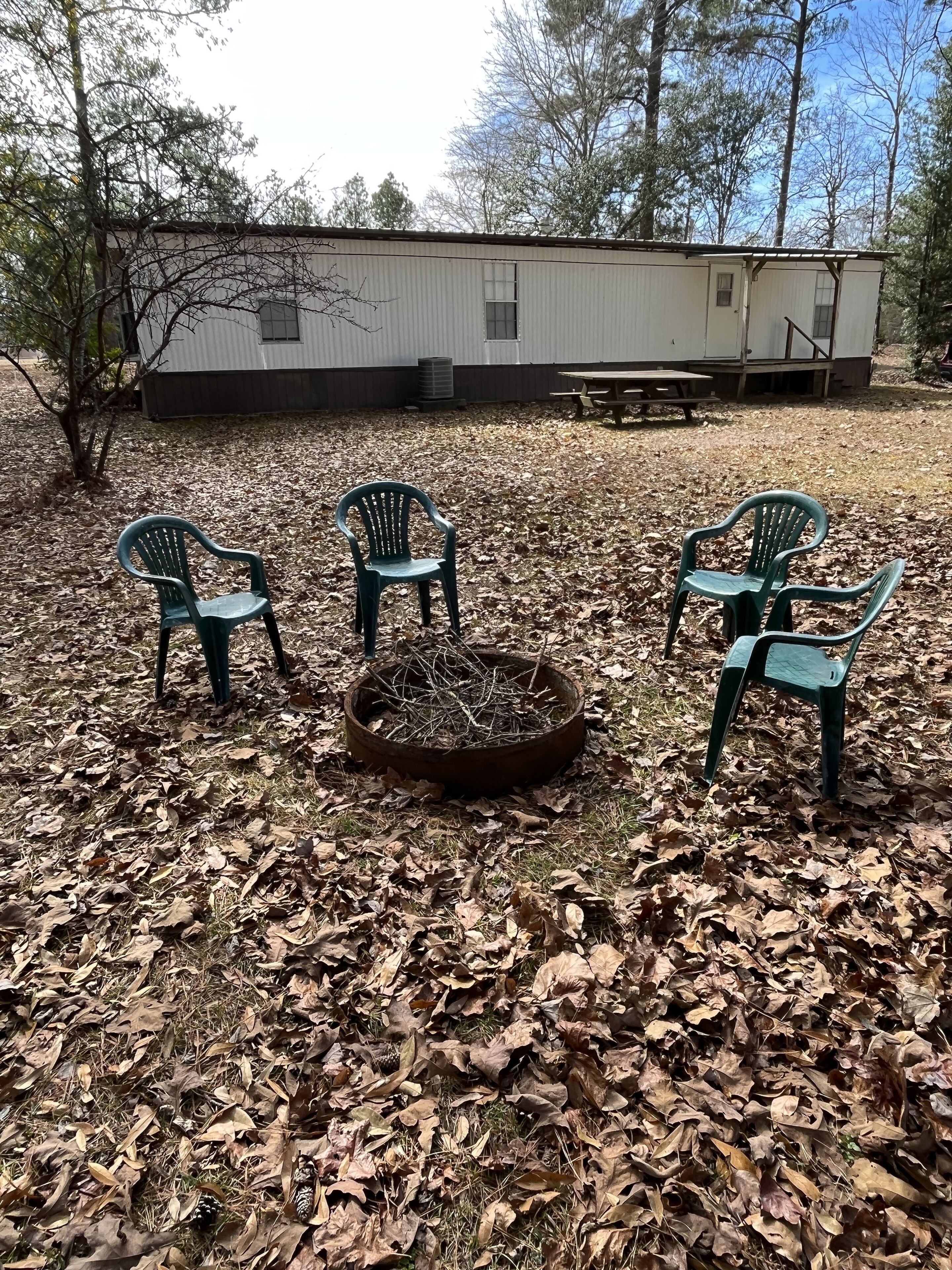 Een achtertuin met een vuurkorf, omringd door vier groene plastic stoelen, een picknicktafel en een wit gebouw op de achtergrond.