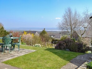 A garden with a metal table and chairs, a stone pathway, and a view of houses and a body of water.