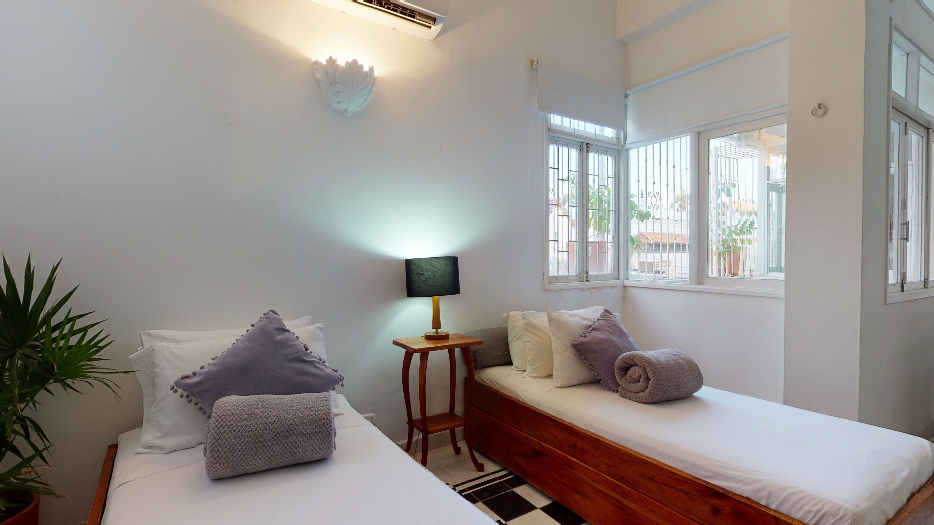 Une chambre avec deux lits, une table de chevet avec une lampe, une plante en pot et une vue sur l’extérieur grâce à de grandes fenêtres.