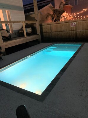 Une piscine rectangulaire dont le bord est recouvert de carrelage, entourée d’une terrasse en bois et d’une clôture.