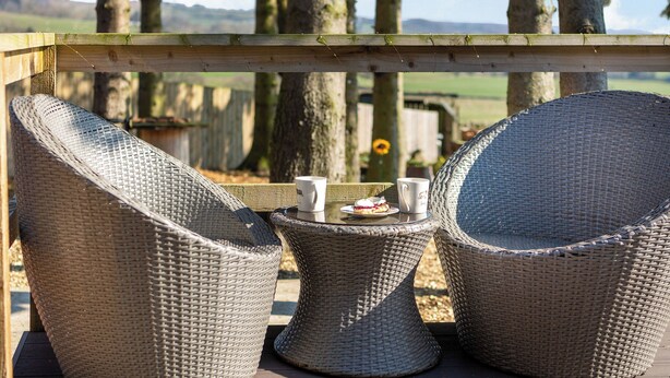 Wicker stoelen en een tafel met een mok en een bord eten op een terras met houten balustrade, met uitzicht op een bebost gebied.
