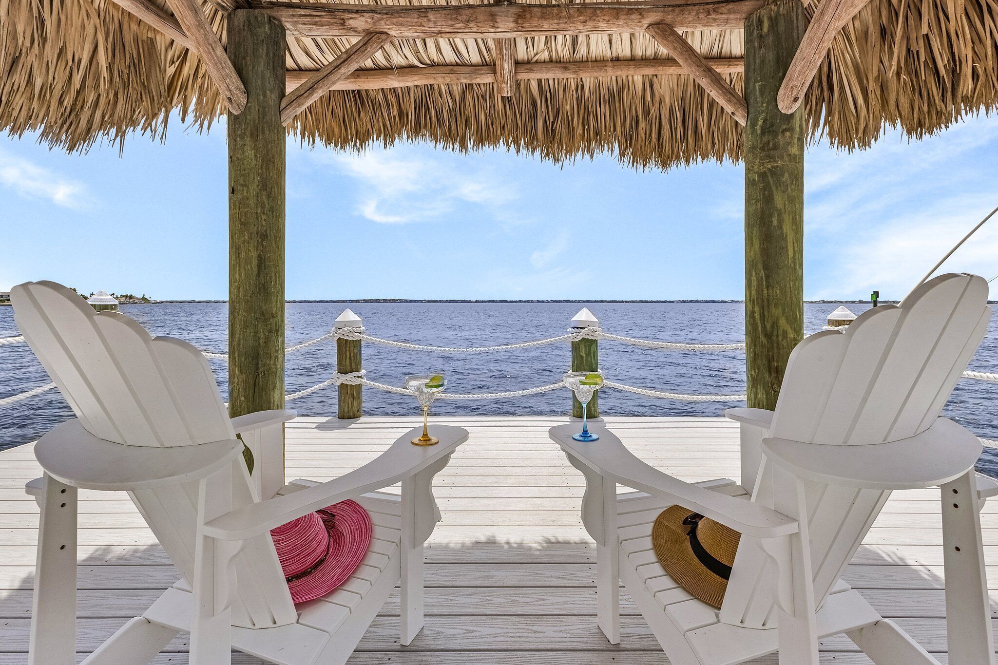 Two white Adirondack chairs with drinks on a wooden deck overlooking the ocean.