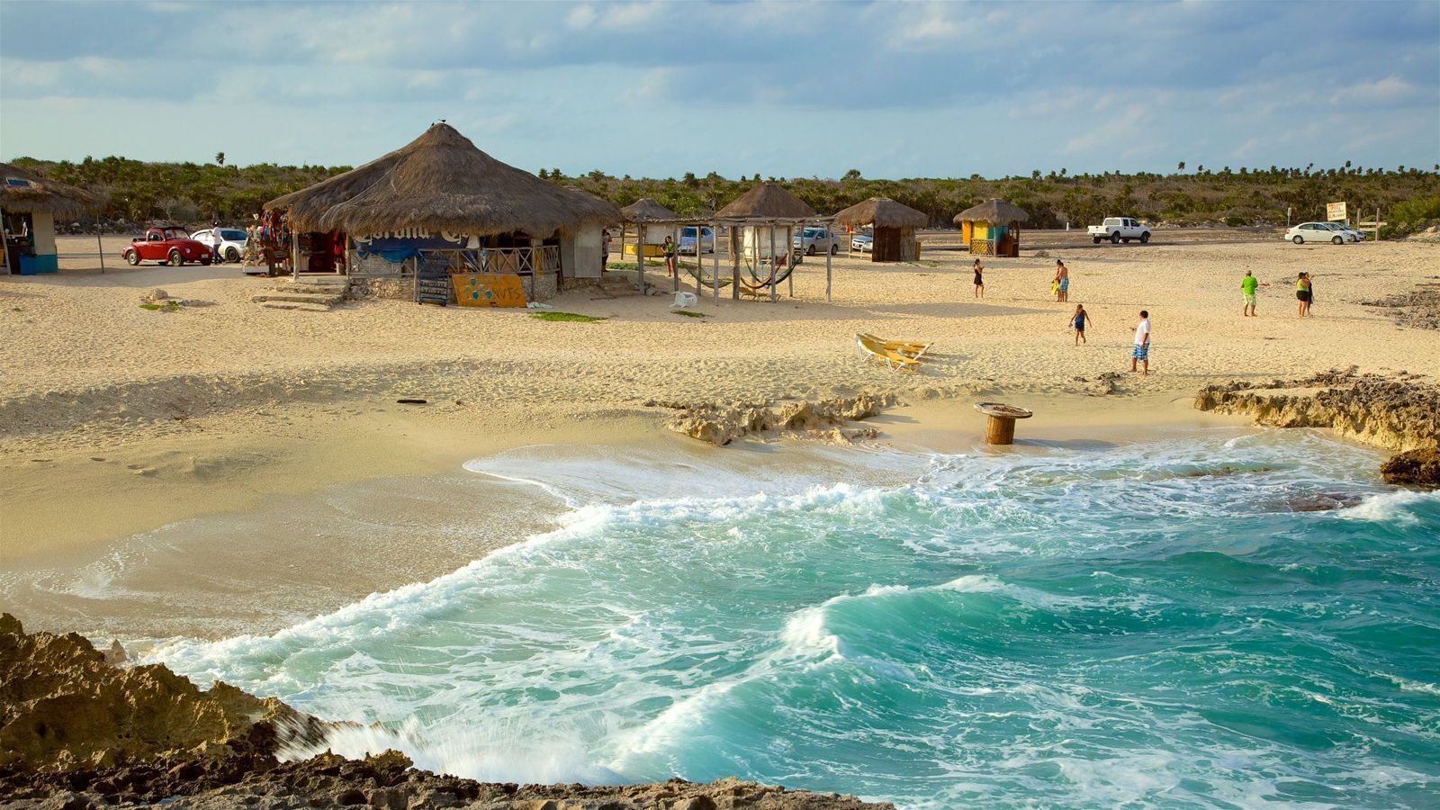 A beach with thatched-roof huts, a rocky shoreline, and a few people walking.