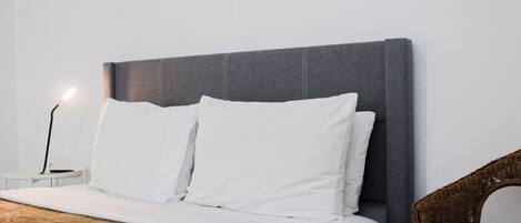 Un lit bien fait, avec du linge de lit blanc et des oreillers, une table de chevet moderne avec une lampe, et une chaise en osier.