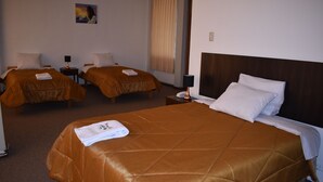 Habitación de hotel con dos camas individuales y una cama doble, cada una con cubrecama marrón y almohadas blancas. Hay una mesita con lámpara entre las camas, y un cuadro en la pared sobre las camas individuales.