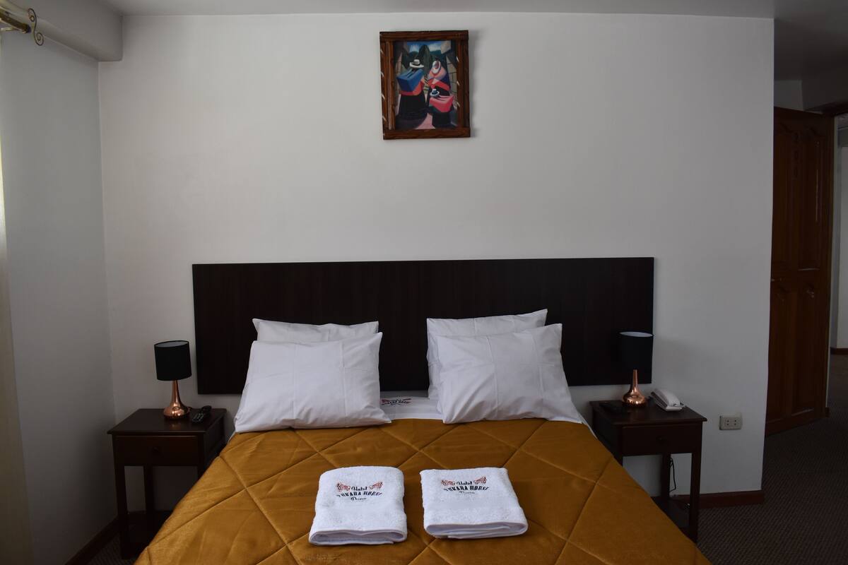 Ett hotellrum med en stor säng, två vita kuddar, en sänggavel, en tavla på väggen och två sängbord med lampor och en telefon.