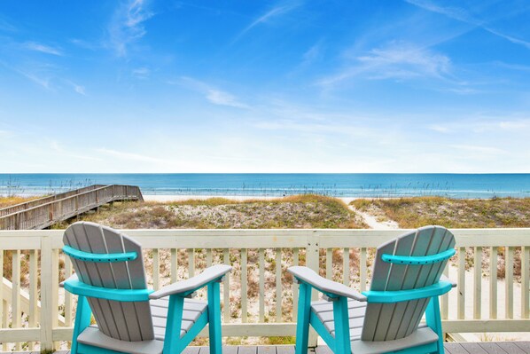 Two Adirondack chairs facing a beach with a wooden boardwalk and dunes.