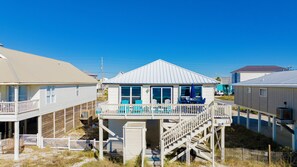 Een huis aan het strand met een balkon, buitenmeubels en een trap naar de bovenverdieping.