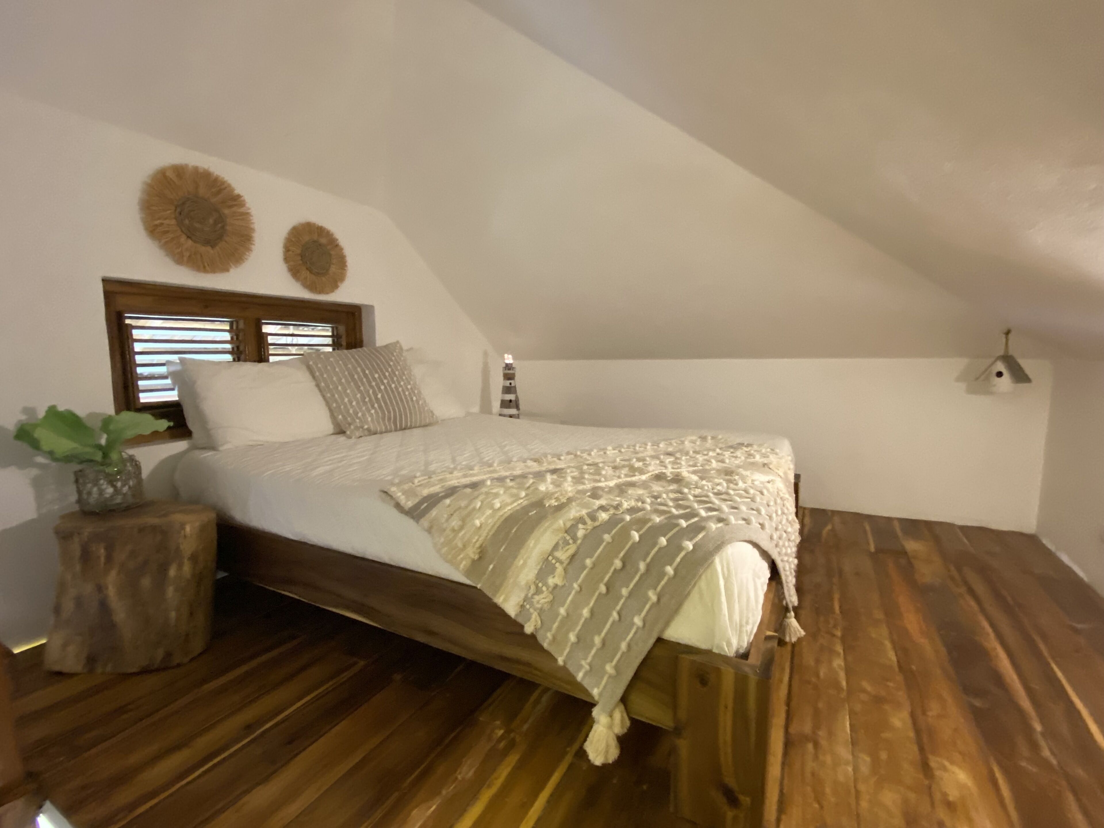 A bedroom with a wooden bed frame, a white bedspread, and a wooden floor. There is a window with blinds, a bedside table with a plant, and a wall-mounted light fixture.