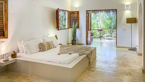 A bedroom with a large bed, a bedside table, a potted plant, and a view of greenery through a window.