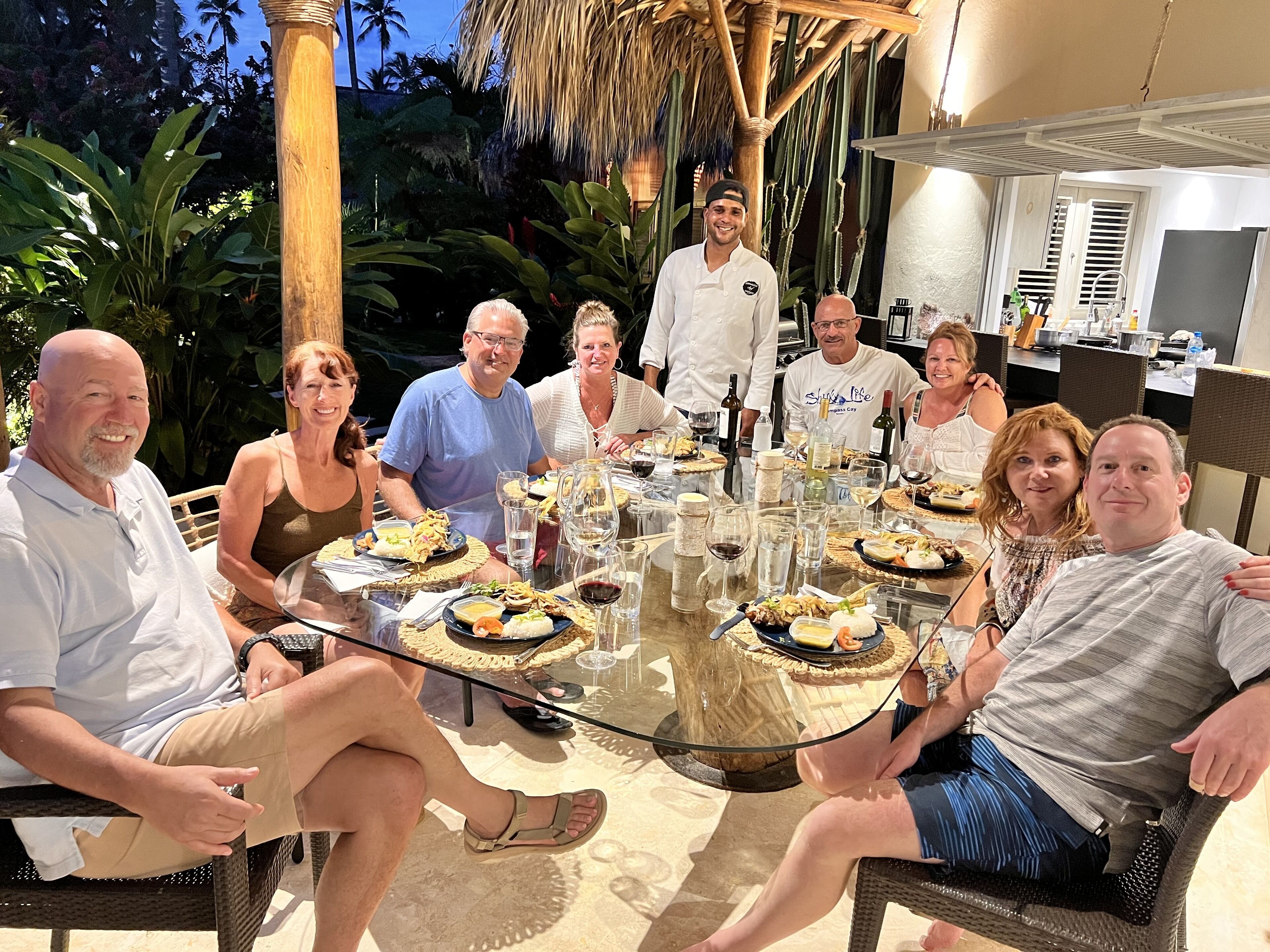 A group of people seated around a round table with various dishes and drinks, outdoors with palm trees and a thatched roof in the background.