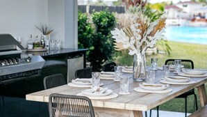 Een buitenterras met een lange houten tafel voor zes personen, omringd door stoelen. Links is een barbecue- en barruimte zichtbaar.