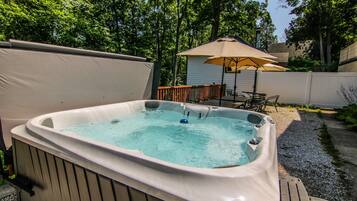 Een hottub in een achtertuin met een houten terras, een patio tafel en een parasol.