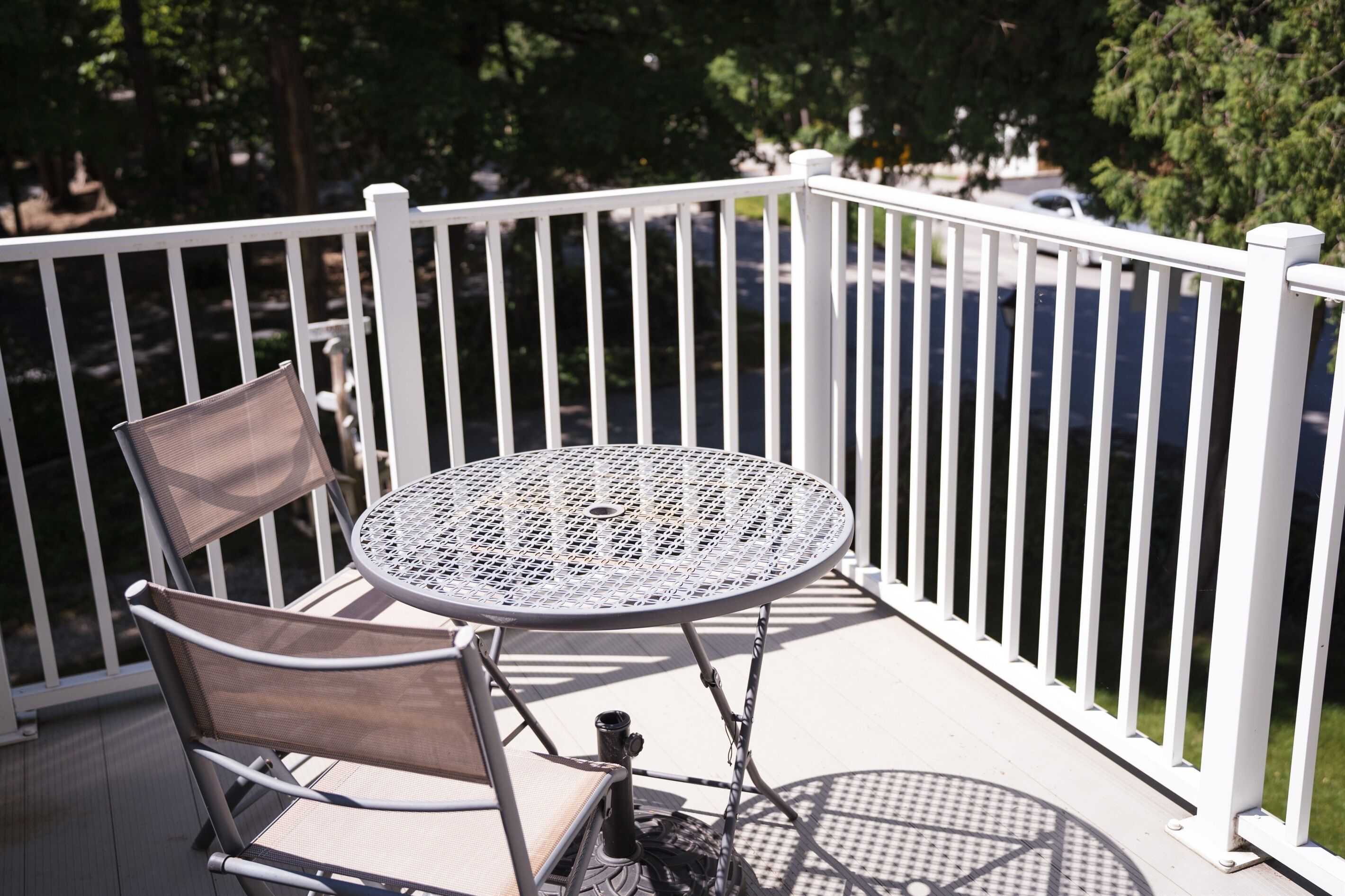 Une table ronde en métal, dotée d’un plateau en treillis métallique, et deux chaises avec des sièges rembourrés, sur un balcon avec des balustrades blanches.