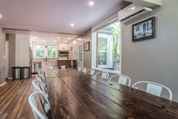 A spacious dining area with a long wooden table, white chairs, and a kitchen in the background.