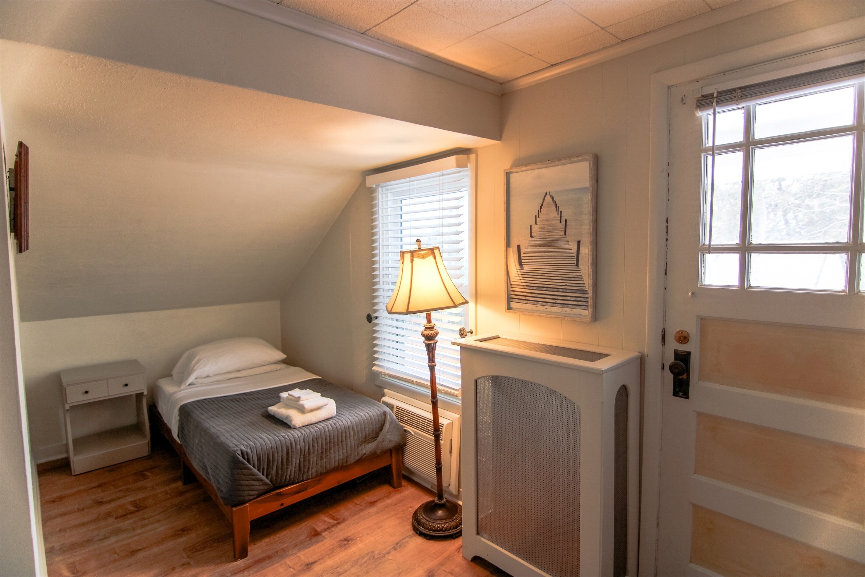 A bedroom with a bed, bedside table, lamp, radiator, and a door with a window.