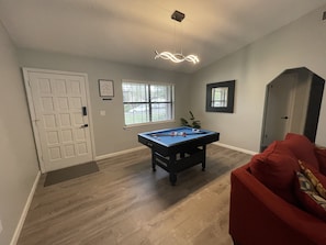 Une pièce avec une table de billard, un canapé rouge, une porte et une fenêtre avec des stores.