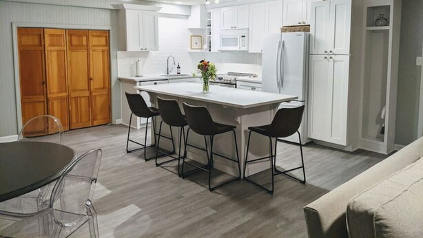 A modern kitchen with white cabinets, a central island with bar stools, a dining table with chairs, and a wooden door in the background. - Beautifully Renovated Space in Lawrenceville Home, WiFi, Smart TV in All Rooms! (Lawrenceville)
