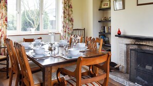 Un comedor con una mesa de madera dispuesta para cuatro, rodeada de sillas, una chimenea y una estantería con libros y objetos decorativos.