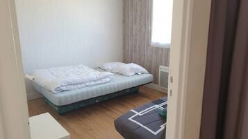 A small, neatly made bed with white bedding in a compact room with wooden flooring and a window with curtains.