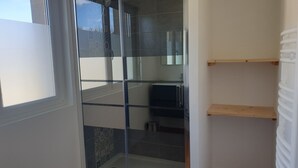 A bathroom with a glass shower enclosure, a toilet, and a sink. There is a window providing natural light, and a wooden shelf is visible. - House equipped for 1-8 people (modular) (Montfarville)