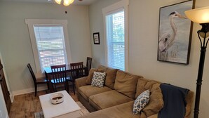 Ein Wohnzimmer mit einem braunen Sofa, einem weißen Couchtisch und einem Esstisch mit Stühlen. Eine Lampe und ein gerahmtes Bild sind an der Wand zu sehen.