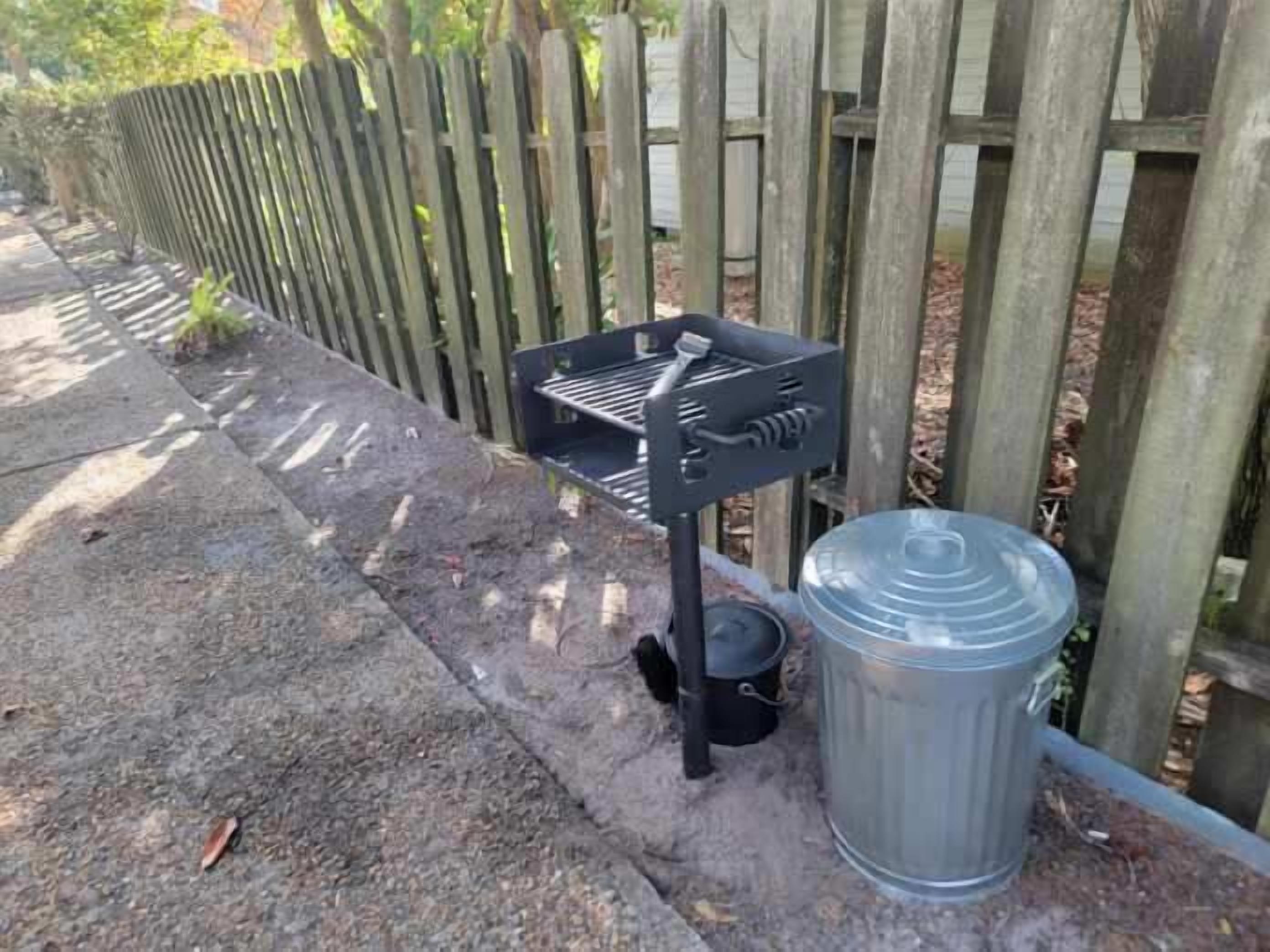 A portable grill with a lid and a small pot on a concrete surface next to a wooden fence.
