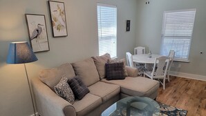 Un soggiorno con un divano angolare beige, un tavolino in vetro e un tappeto decorato. Si notano due opere d'arte appese alle pareti, una lampada e un'area pranzo con un tavolo e sedie bianche.