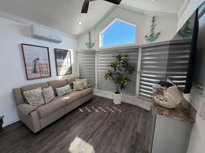 A living room with a beige sofa, a potted plant, and a large window with blinds.